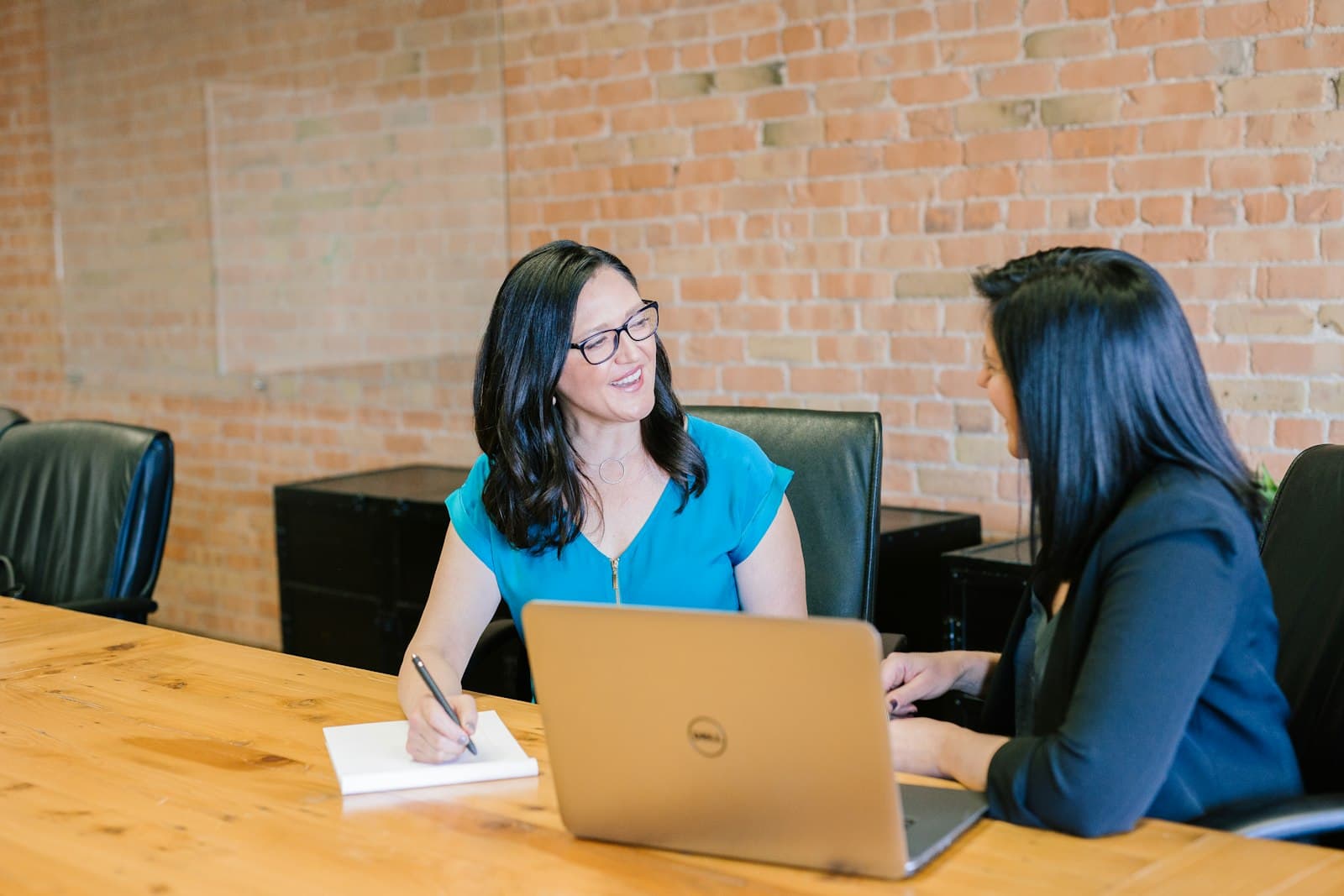 Two colleagues talking through a project at a desk while taking notes on a laptop